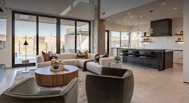 Modern open-concept living room and kitchen with curved seating, round wood table, and floor-to-ceiling windows overlooking a